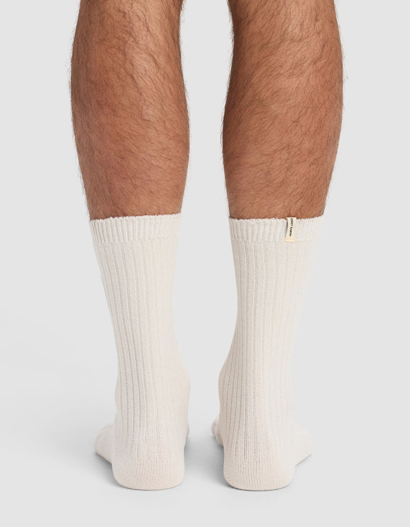 Back view of a person's lower legs wearing The Plush Lounge Calf Sock 3-Pack by Cozy Earth—thick, ribbed, off-white socks with a small beige tag near the top edge. Plain, light-colored background.