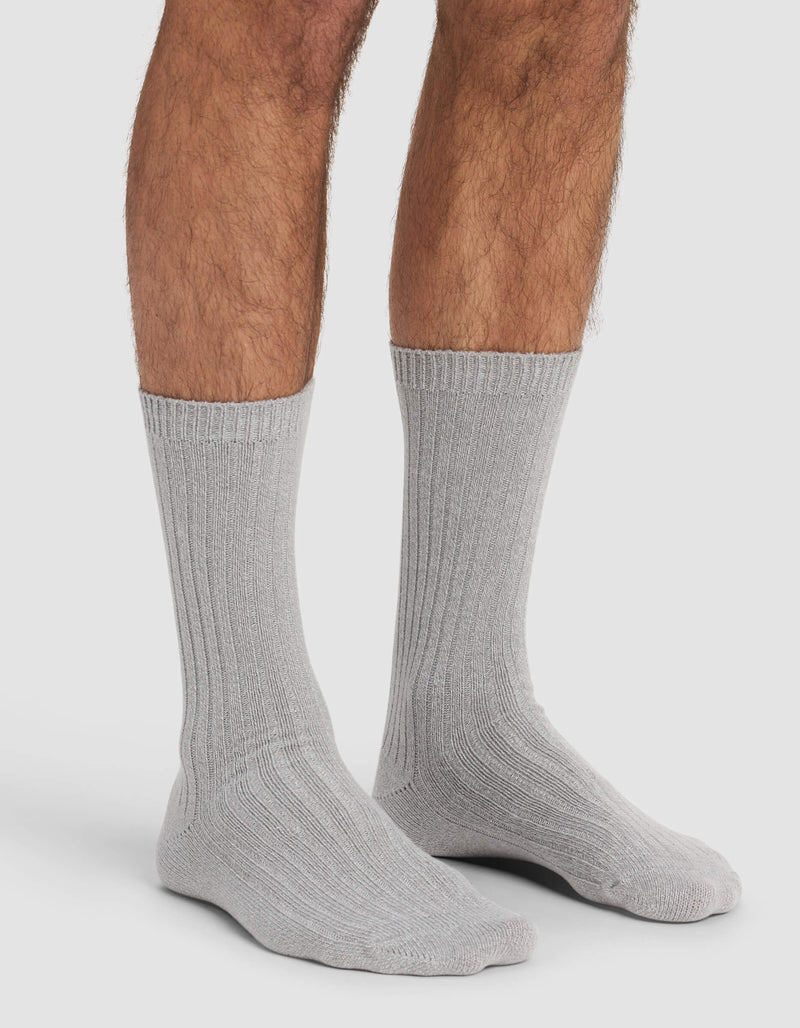 A person wears Cozy Earth's Men's Plush Lounge Calf Socks in light gray, standing on a white background with only their lower legs and feet visible.