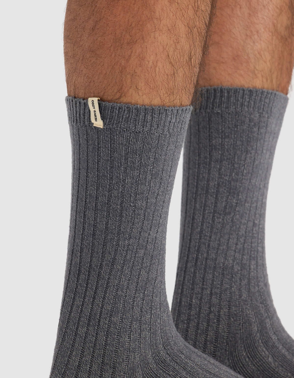 Close-up of a person's lower legs wearing The Plush Lounge Calf Sock in dark gray from Cozy Earth, featuring ribbed texture and a small beige tag near the top. Plain white background.