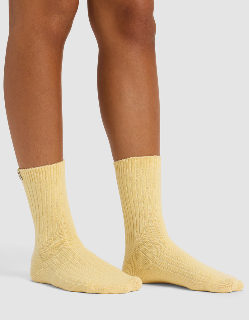 Someone wearing The Plush Modern Crew Sock by Cozy Earth in light yellow stands on a pale surface, with only their lower legs and feet visible.