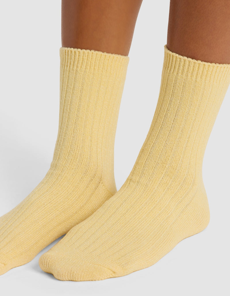 A close-up of two feet wearing Cozy Earth’s Plush Lounge Quarter Socks in light yellow, covering the ankles and lower calves, set against a plain light gray background.