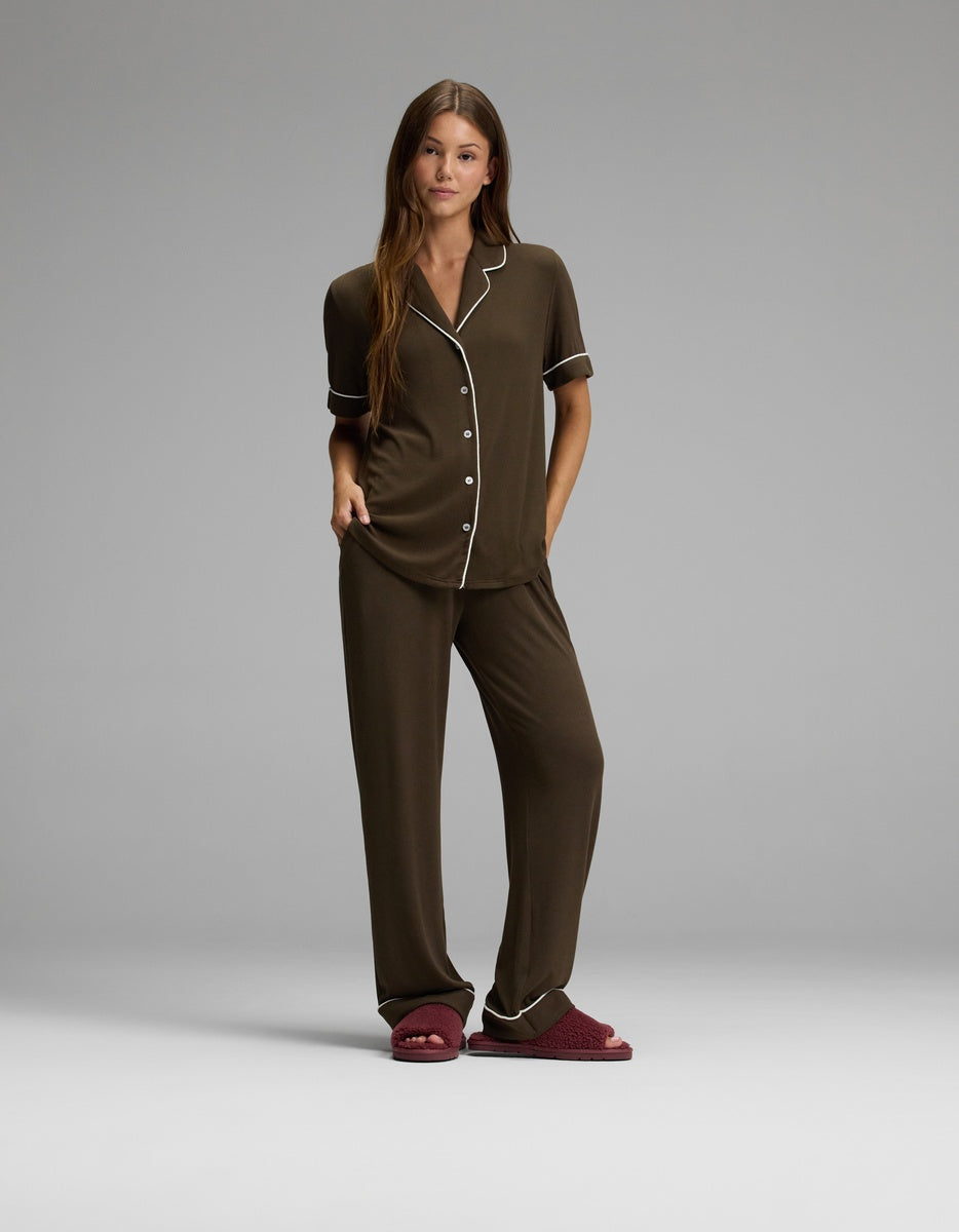 A woman stands wearing brown pajamas with white piping and Cozy Earth Puffy Sheep Slides. She has long brown hair and a relaxed posture against a plain gray background.