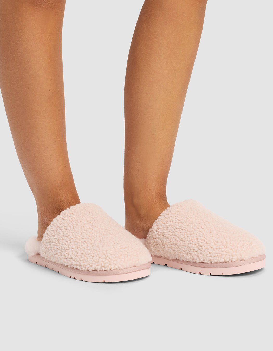 A person wears Cozy Earth Puffy Sheep Slippers in light pink, featuring a soft, sherpa-like texture and flat sole, while standing on a light gray surface. Only their lower legs and feet are visible.|Color:Blossom