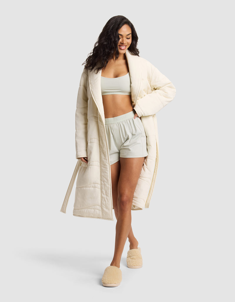 A woman smiles with one hand in her pocket, wearing the Cozy Earth Women's Quilted House Coat over a light sports bra and matching shorts, paired with fuzzy beige slippers, set against a plain light background.