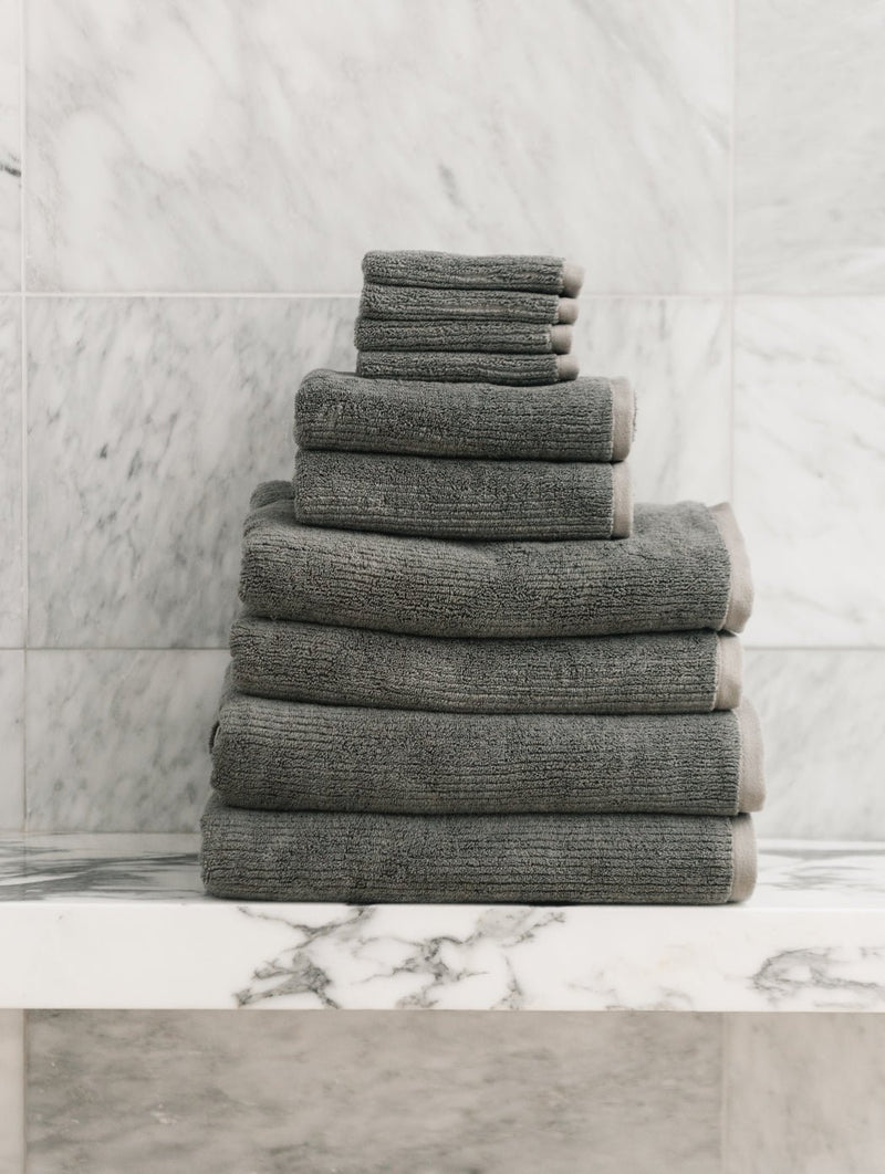 The Cozy Earth Complete Ribbed Terry Bath Bundle, featuring gray towels in assorted sizes, is displayed neatly on a white marble shelf with a marble-tiled wall backdrop.