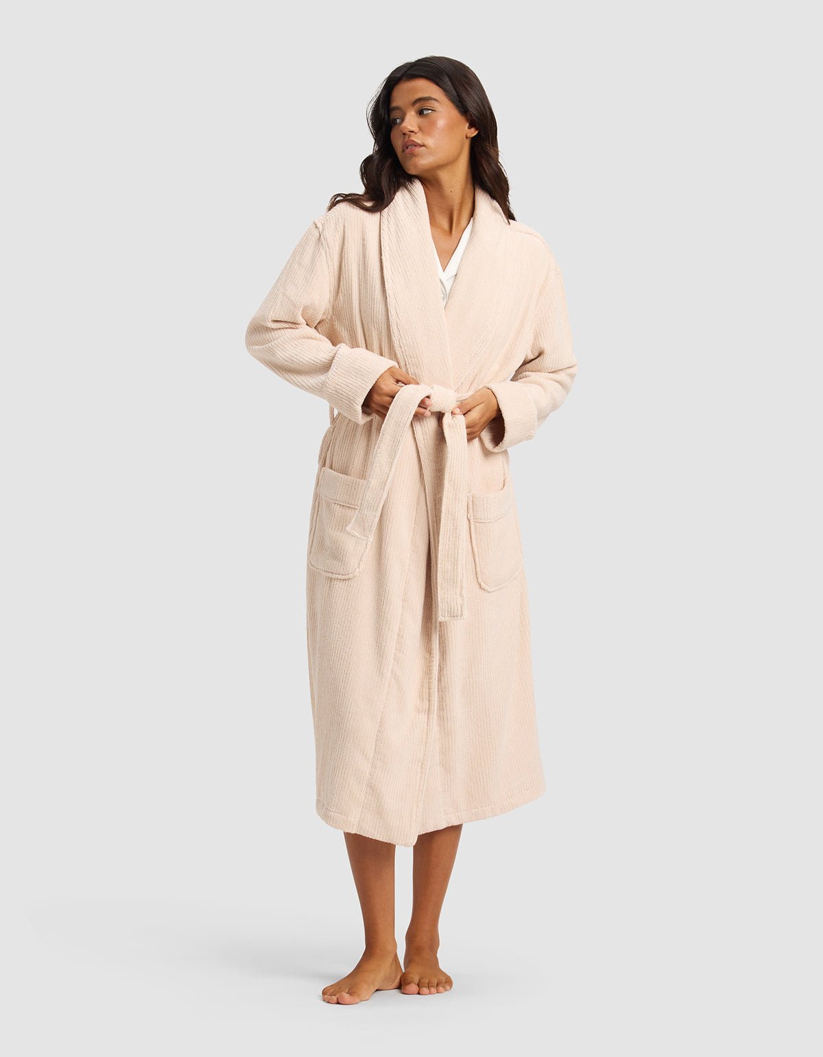 A woman stands barefoot against a plain light gray background, wearing Cozy Earth's Ribbed Terry Bath Robe in light beige featuring long sleeves, large front pockets, and a belt.|Color:Petal