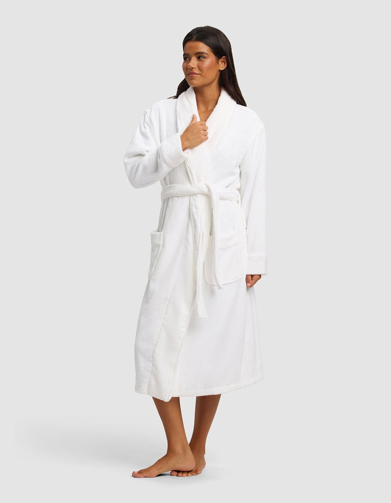 A woman stands barefoot against a light gray backdrop, wearing the Cozy Earth Ribbed Terry Bath Robe with long sleeves, large front pockets, and a belt. She looks to the side while holding the robe closed with one hand.