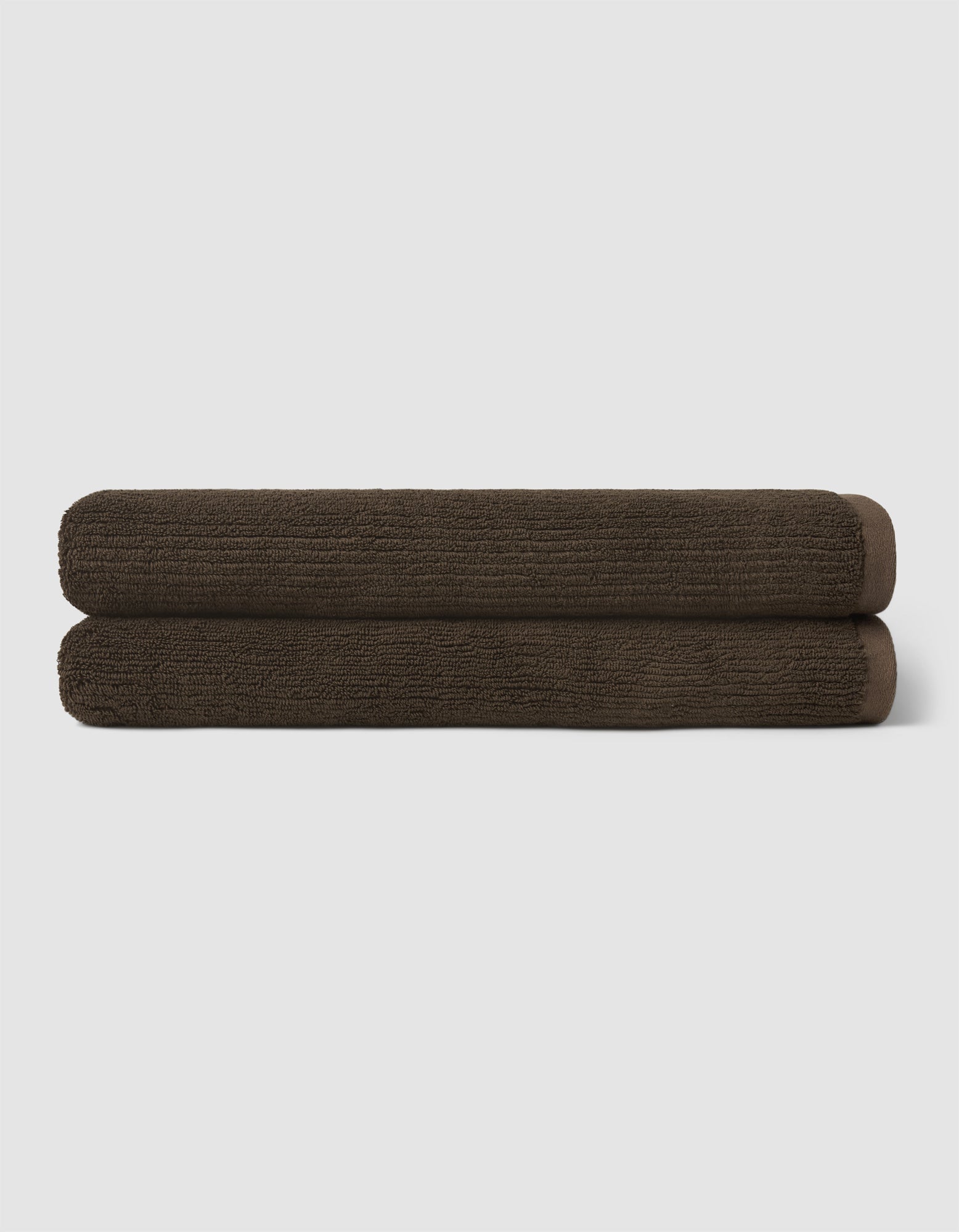 Two dark brown towels from the Cozy Earth Complete Ribbed Terry Bath Bundle are neatly rolled and stacked atop each other against a plain light gray background.|Color:Espresso