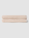 Two neatly folded beige towels from Cozy Earth's Complete Ribbed Terry Bath Bundle are stacked on top of each other against a plain light gray background.|Color:Petal