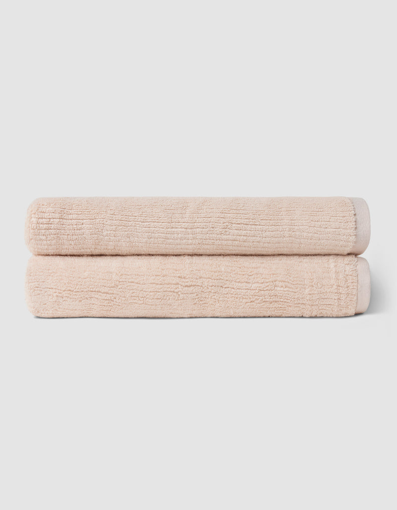 The Complete Ribbed Terry Bath Bundle by Cozy Earth features two neatly rolled, light beige towels stacked on a plain, light gray background.