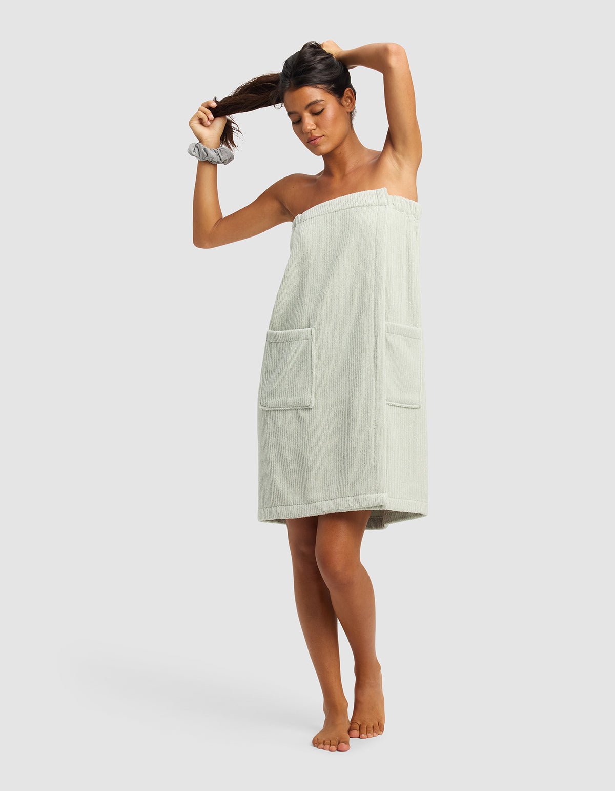 A woman stands barefoot against a plain background, adjusting her ponytail while wearing the Cozy Earth Ribbed Terry Bath Wrap in light green with two front pockets and a matching scrunchie on her wrist.|Color:Laurel