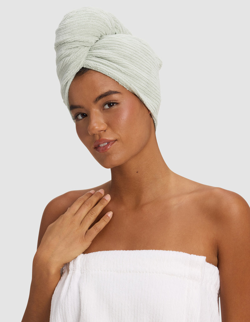 A woman with a Cozy Earth Ribbed Terry Hair Towel in light green wrapped around her head and a white towel around her chest poses against a plain background, gently touching her collarbone and smiling softly.