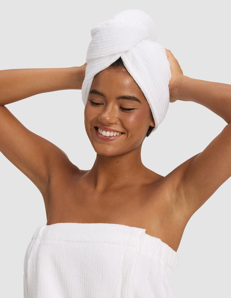 A smiling woman poses with arms raised, wearing a Cozy Earth Ribbed Terry Hair Towel and a white towel wrapped around her body, standing against a plain light gray background.