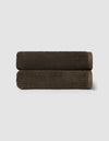 Two neatly rolled, dark brown towels from the Cozy Earth Complete Ribbed Terry Bath Bundle are stacked against a plain light gray background.|Color:Espresso