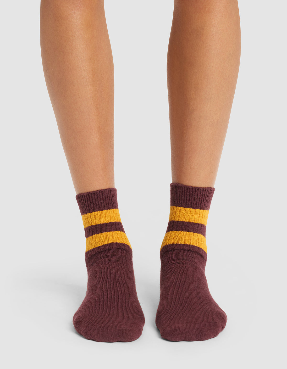 A pair of feet wearing Cozy Earth Women's Sadie Socks, maroon with two yellow stripes at the top, standing on a white background.  |Color:Burgundy/Goldenrod