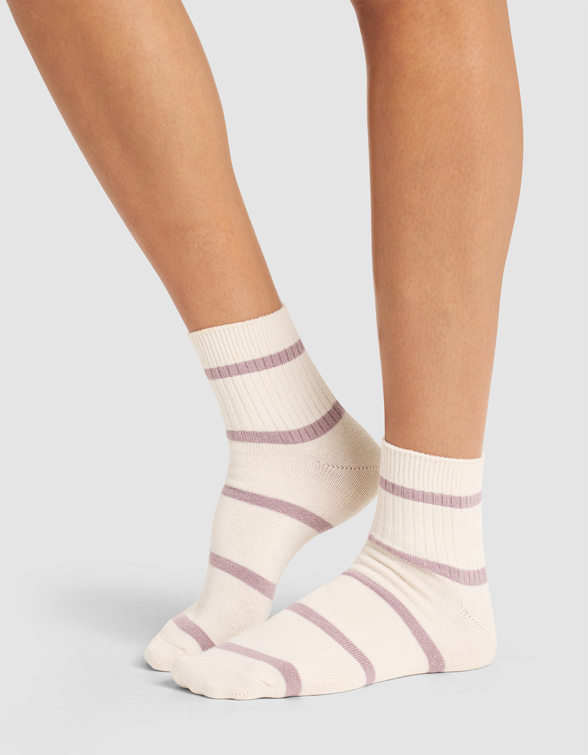 A person stands on tiptoes against a plain light gray background, wearing Cozy Earth's Women's Sadie Sock in cream with light purple horizontal stripes. |Color:Parchment/Dusty Orchid