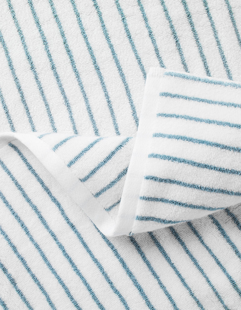 The Cozy Earth Striped Luxe Bath Towel is white with evenly spaced thin blue stripes, shown folded at one corner to reveal both sides.