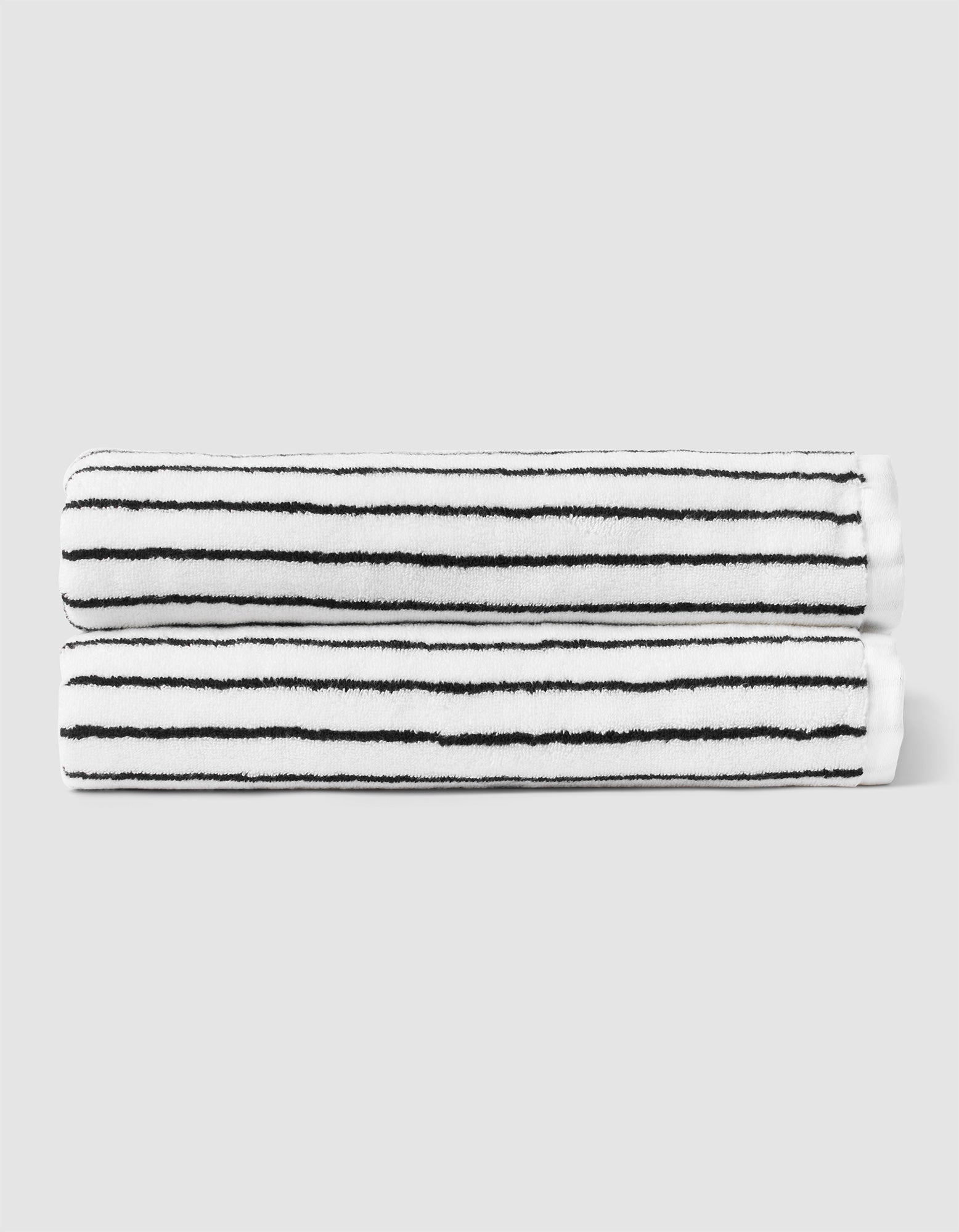 Two Striped Luxe Bath Sheets by Cozy Earth, featuring thin black horizontal stripes on white, are neatly rolled and stacked against a plain light gray background. |Color:Black Stripe