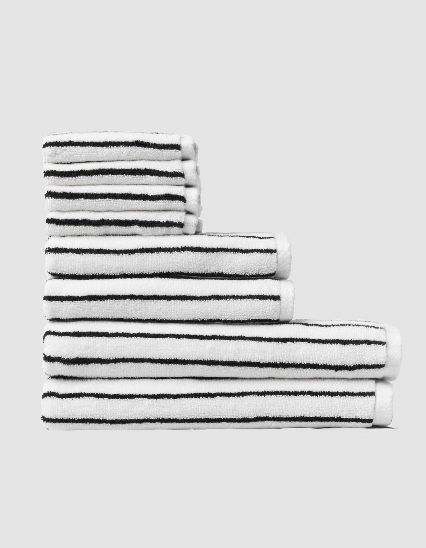 The Cozy Earth Striped Luxe Bath Towel Set features five white towels with thin black horizontal stripes, stacked in ascending order against a plain light gray background. |Color:Black Stripe