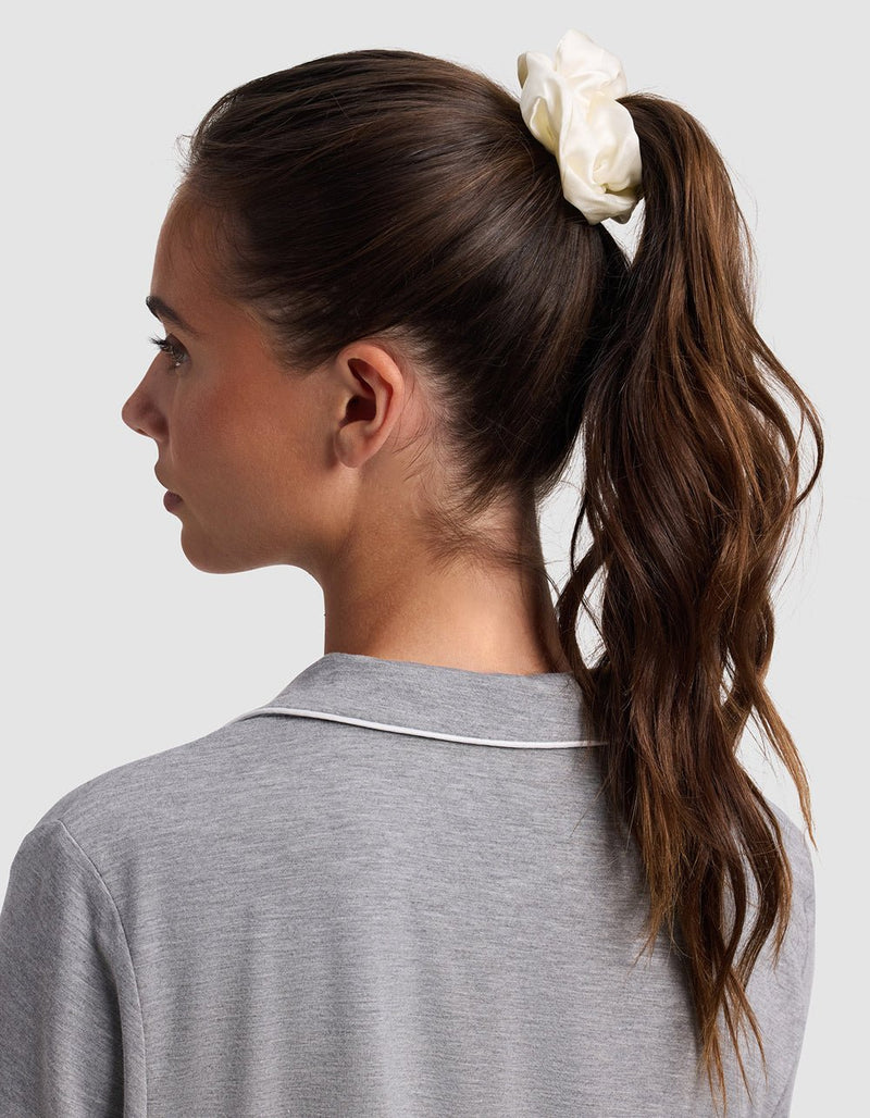 A woman with long brown hair in a high ponytail, secured with a white Cozy Earth Silk Scrunchie – Last Chance, is shown from the side and back. She wears a grey top with white piping on the collar against a plain light background.