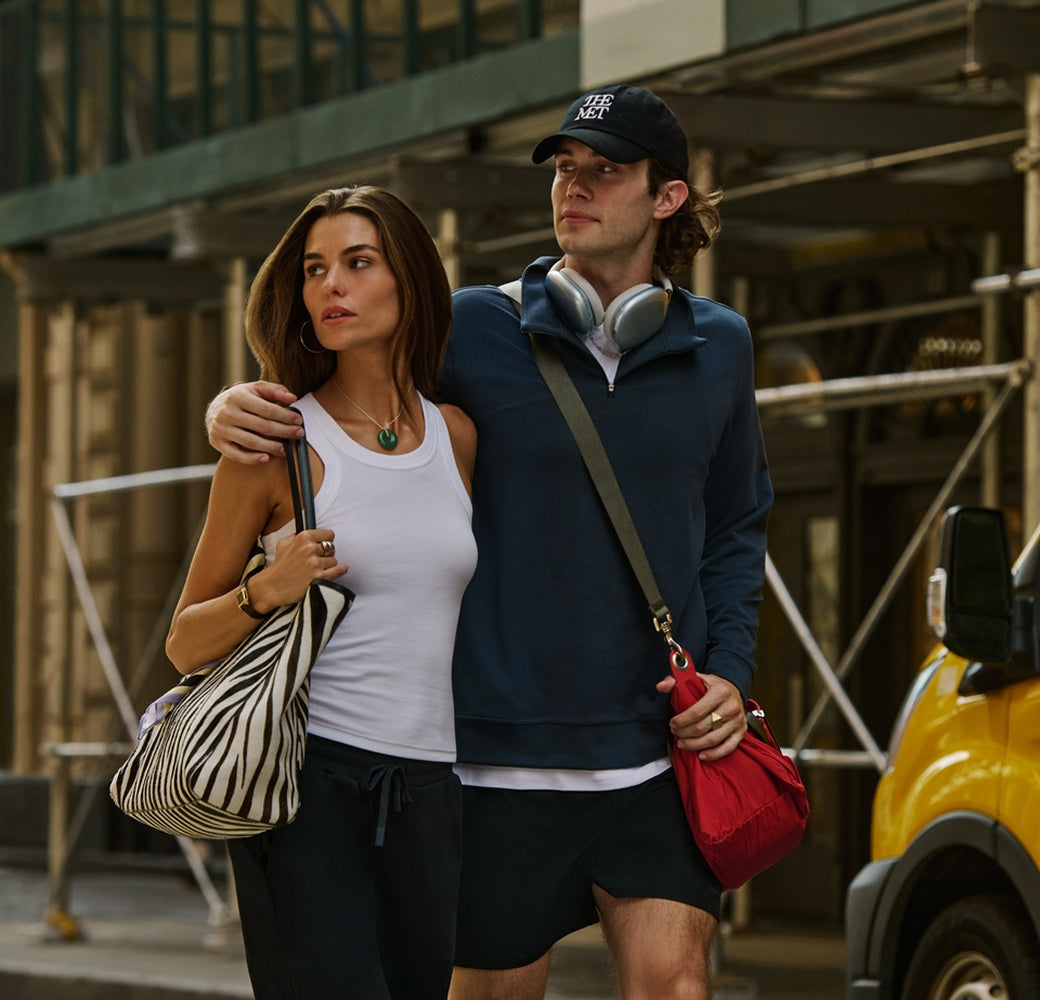 A man and a woman walking arm-in-arm on a city street. The man wears a navy blue quarter-zip hoodie, black shorts, a black baseball cap, and has silver headphones around his neck, with a red crossbody bag hanging from his shoulder.
