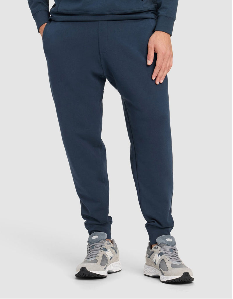 A person wears Cozy Earth's Men's StretchTech Quarter Zip & Jogger Set in navy, paired with light gray sneakers, standing with one hand in their pocket against a plain light background.A man stands against a plain background wearing Cozy Earth’s Men's StretchTech Jogger in black, paired with a black sweatshirt and gray slippers. He has one hand in his pocket and looks slightly to the side.