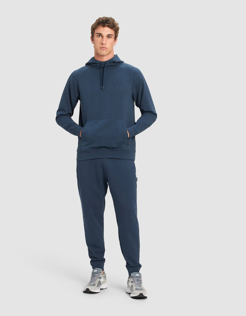 A young man stands facing forward, wearing a Cozy Earth Men's StretchTech Jogger set in dark blue with gray sneakers, hands in his hoodie pocket, against a plain light gray background.