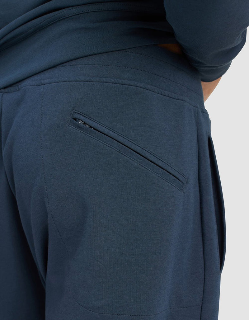 Close-up of a person wearing Cozy Earth Men's StretchTech Quarter Zip & Jogger Set in dark blue, showing a zippered back pocket and one hand partly in the waistband; plain light gray background.