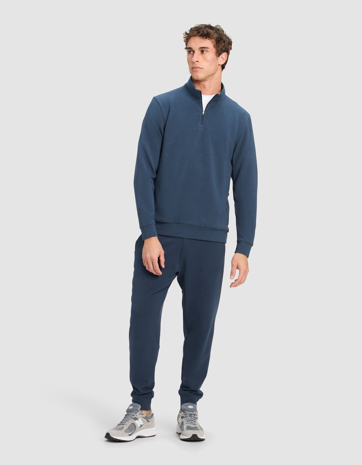 A man wears Cozy Earth's Men's StretchTech Quarter Zip and matching jogger pants with light gray sneakers, standing slightly turned with a neutral expression against a plain light background.