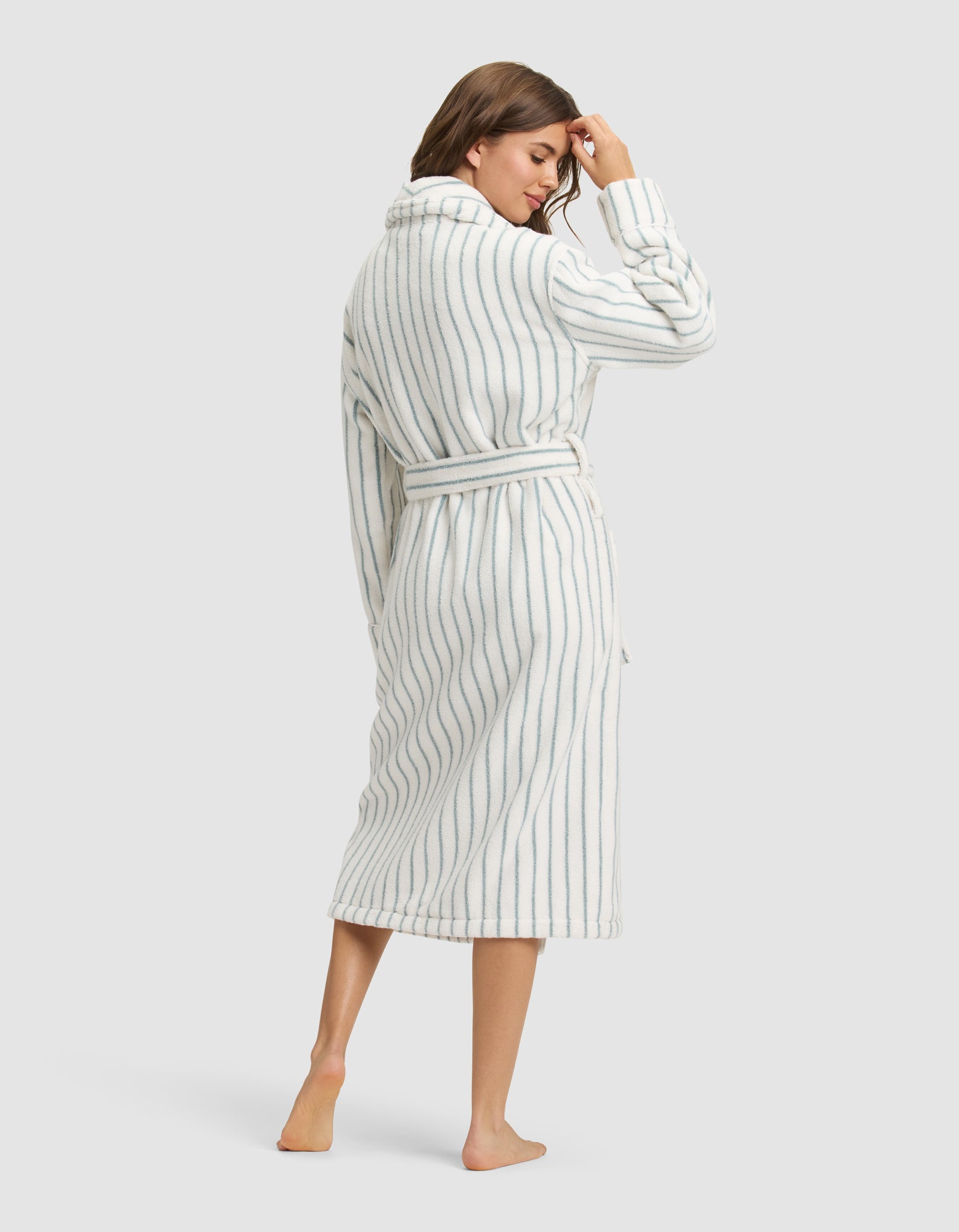 A woman stands barefoot, facing away, wearing the Cozy Earth Striped Luxe Bath Robe—a long, belted white robe with vertical blue stripes—touching her hair against a plain light background. 