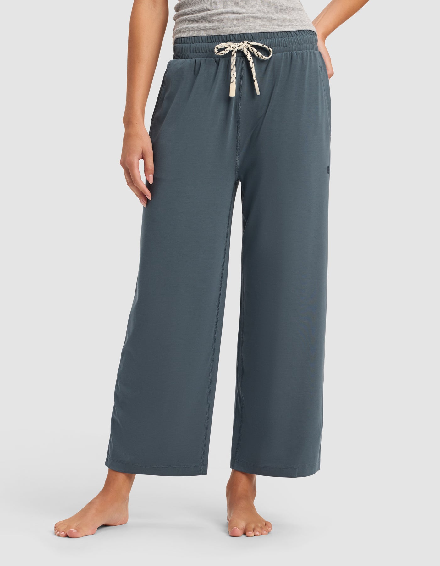 A person stands barefoot wearing Cozy Earth’s Women's Studio Cropped Wide Leg Pant in blue-gray with an elastic waistband and striped drawstring, paired with a gray top; their left hand rests on their hip.