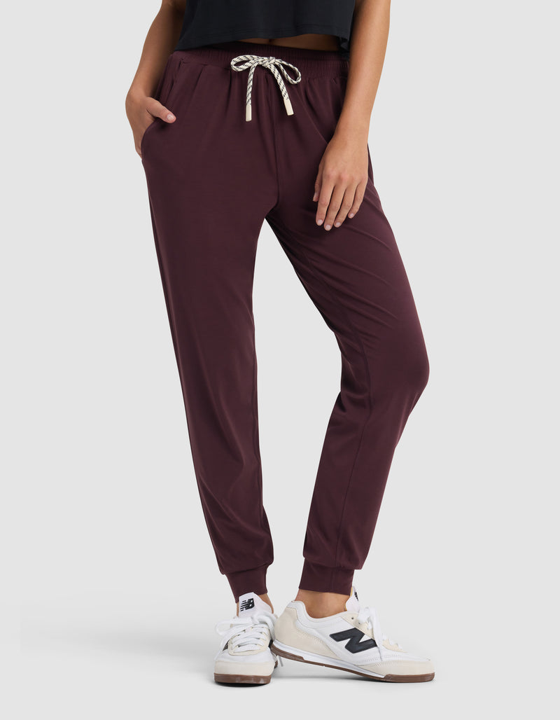 A person stands with hands in pockets against a plain light background, wearing Cozy Earth Women's Studio Jogger pants in maroon, a black top, and white New Balance sneakers.