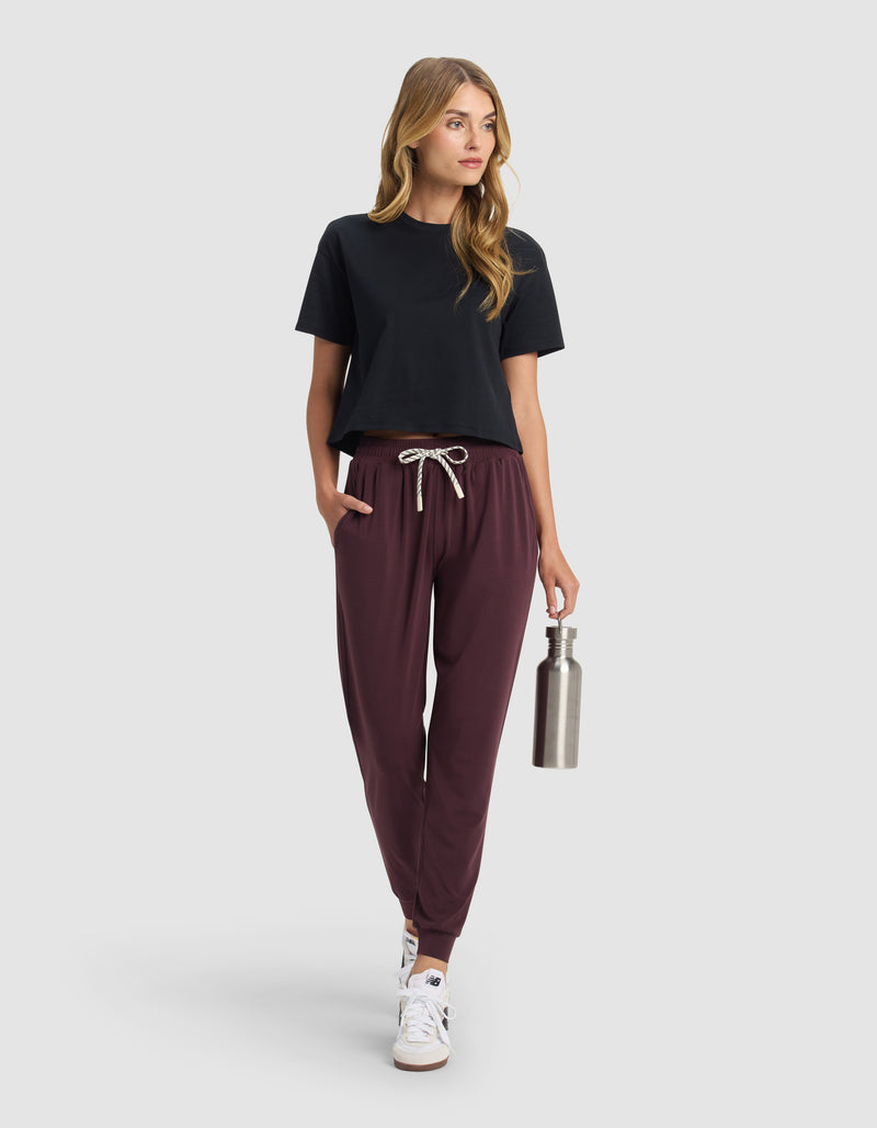 A woman with long hair models Cozy Earth's Women's Studio Jogger in maroon, paired with a black t-shirt and white sneakers. She holds a stainless steel water bottle and looks to the side against a plain light gray background.