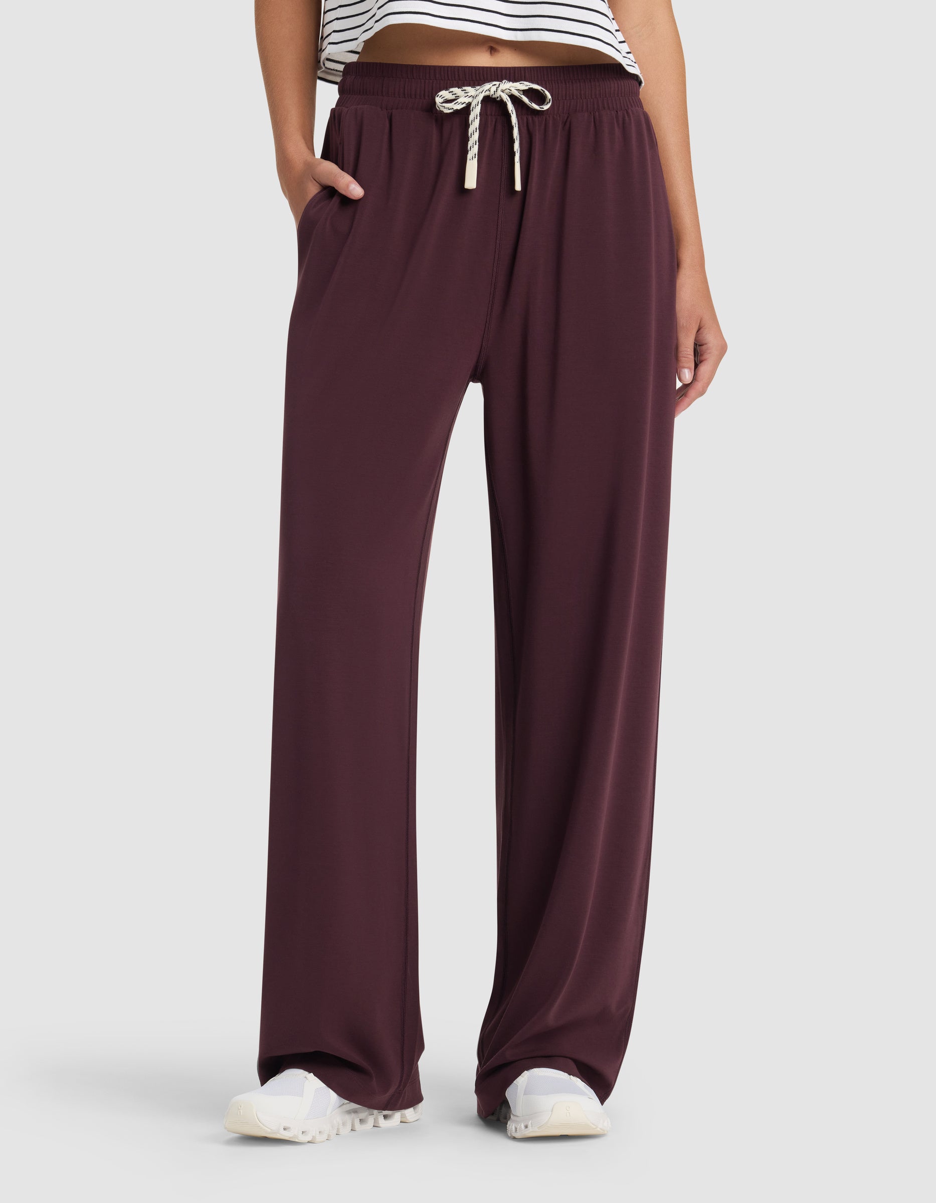 A person wears Cozy Earth Women's Studio Wide Leg Pant in burgundy with a white drawstring, white sneakers, and a cropped striped top. Their left hand is in their pocket; only the lower body is shown. 