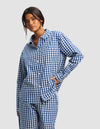 A woman with dark hair models the Women's Sutton Long Sleeve Pajama Top by Cozy Earth in blue and white gingham, standing against a light background with one hand on her hip.|Color:Pacific Gingham