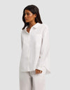 A woman with long dark hair wears the Cozy Earth Women's Sutton Long Sleeve Pajama Top, a loose-fitting white button-up with a chest pocket, paired with matching white pants, against a plain light gray background.|Color:White