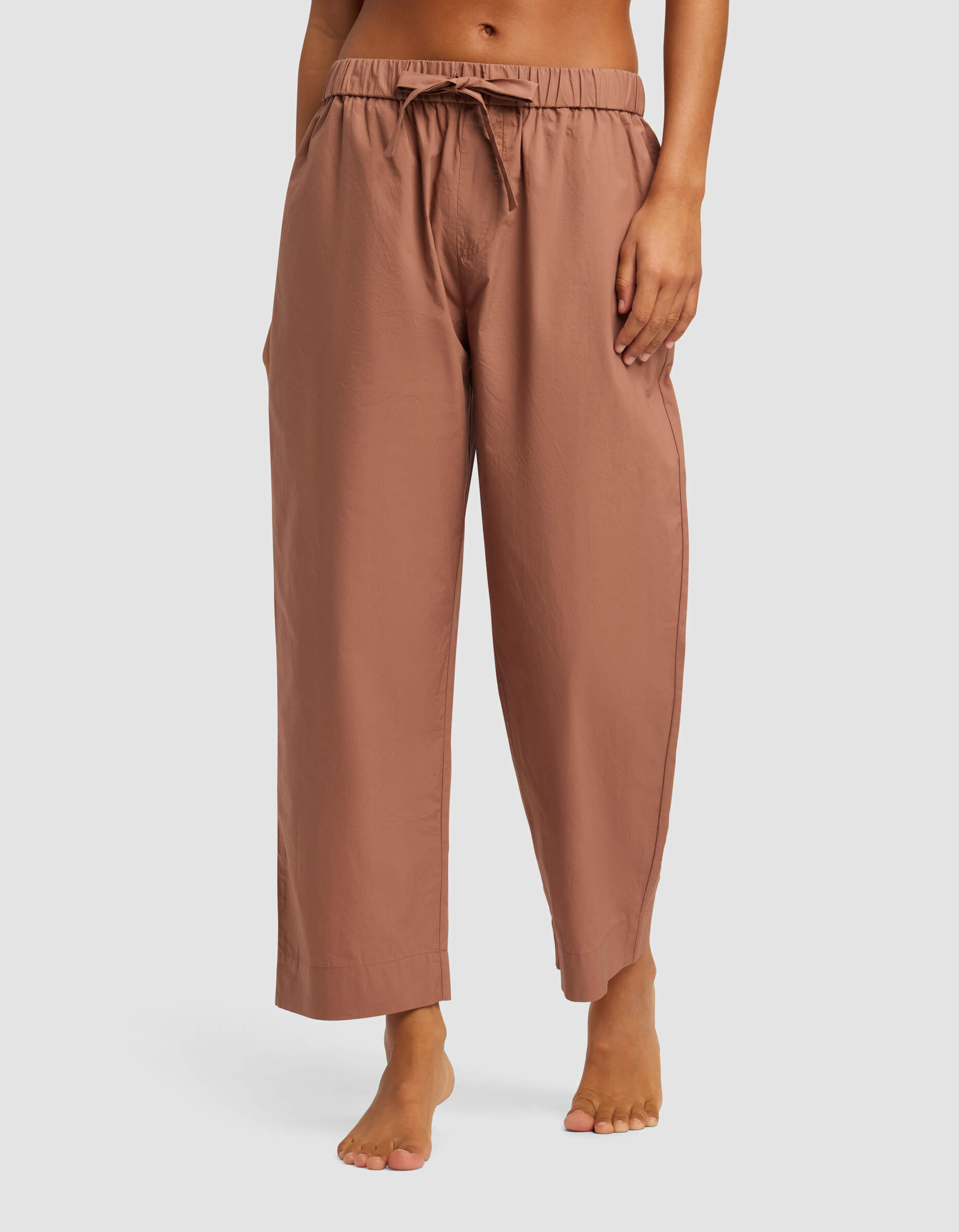 A woman stands barefoot in Cozy Earth’s Women’s Sutton Cropped Pajama Pant—loose, light brown drawstring pants with side pockets, cropped above the ankles—against a plain light background.
