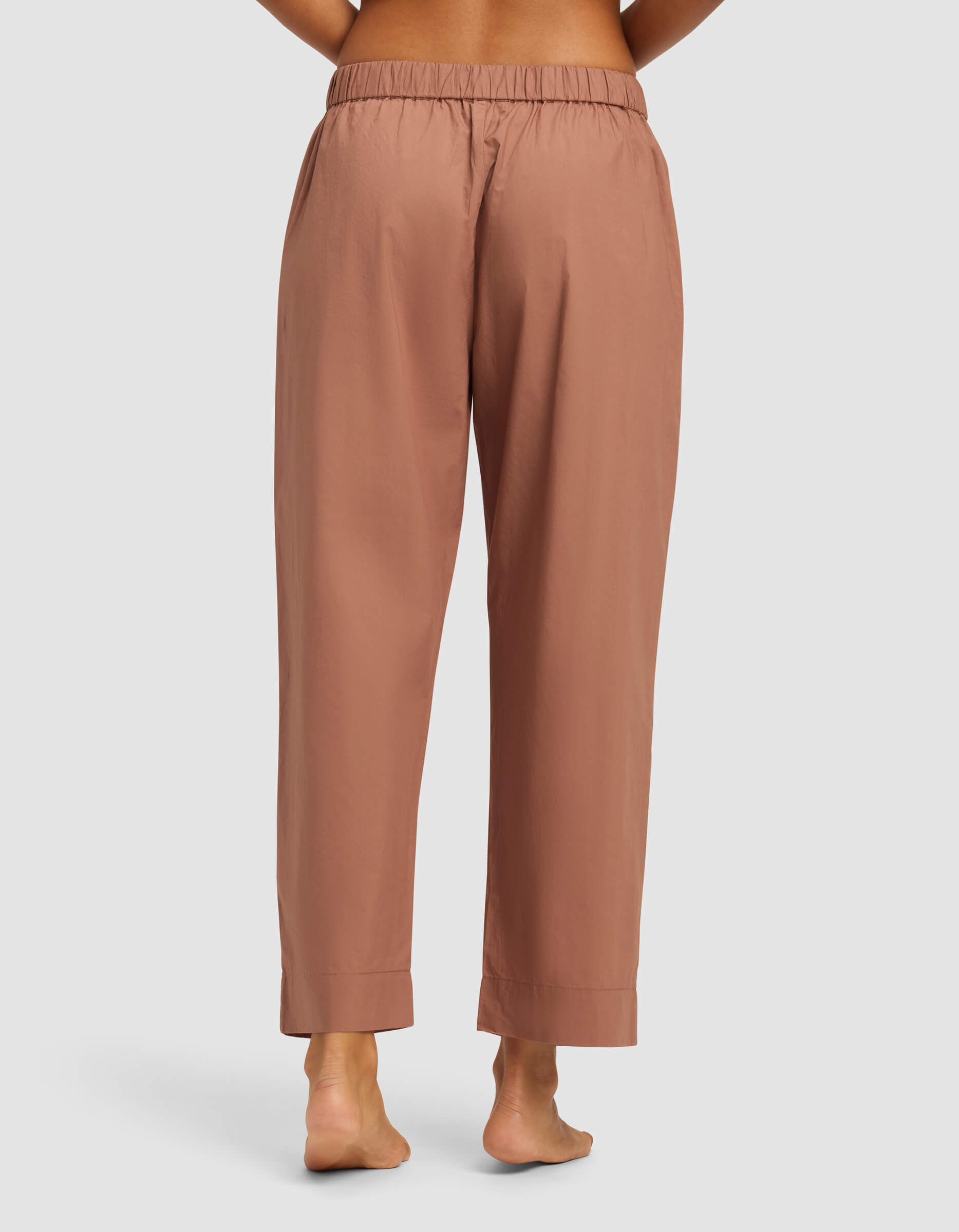 A person is barefoot on a light surface, shown from the back, wearing Cozy Earth’s Women's Sutton Cropped Pajama Pant—loose-fitting, high-waisted pajama pants in light brown or terracotta with an elastic waistband and relaxed straight legs.