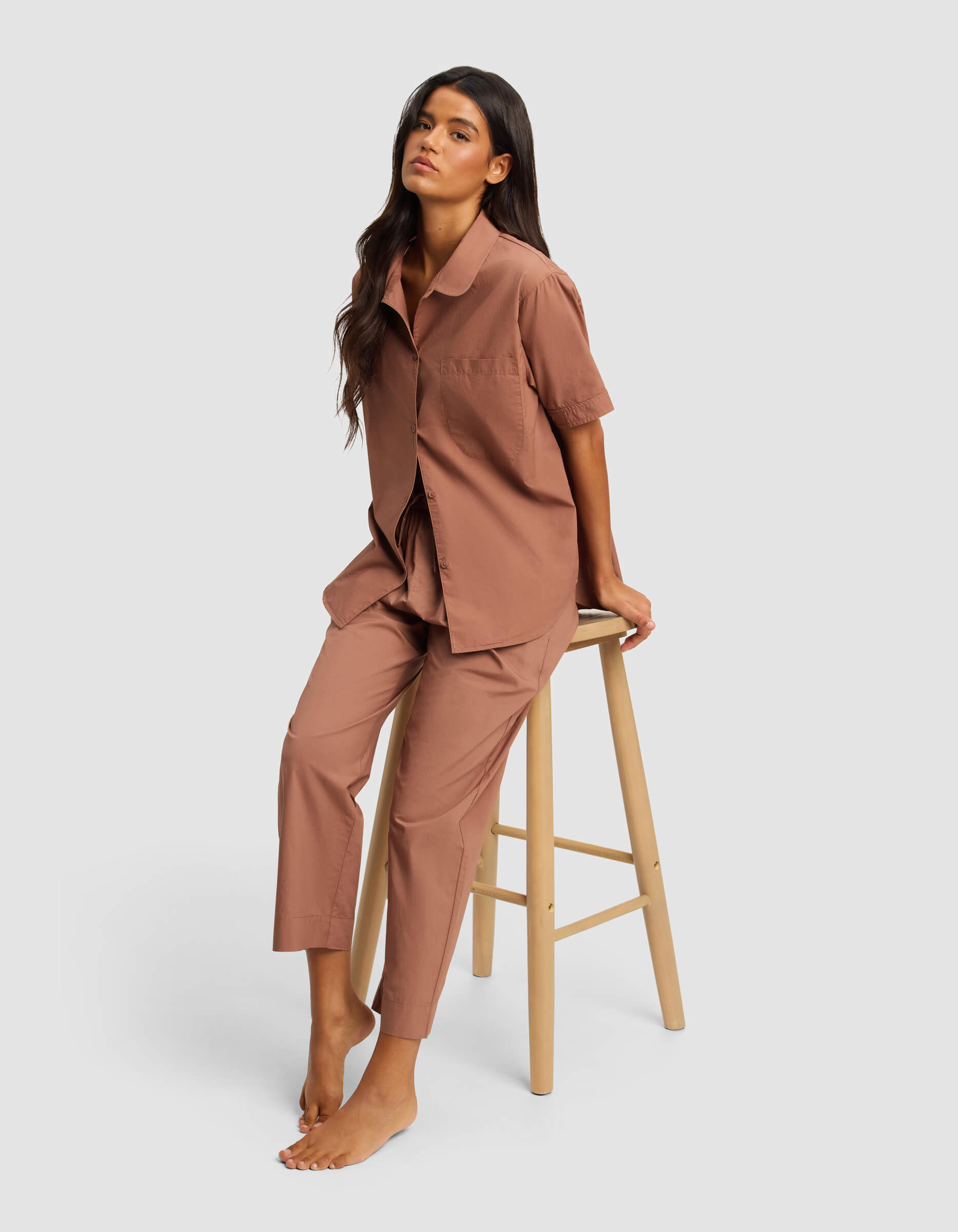 A woman with long dark hair sits barefoot on a wooden stool, wearing Cozy Earth's Women's Sutton Cropped Pajama Pant set in light brown, featuring a matching short-sleeve button-up shirt and pants, against a plain light gray background.