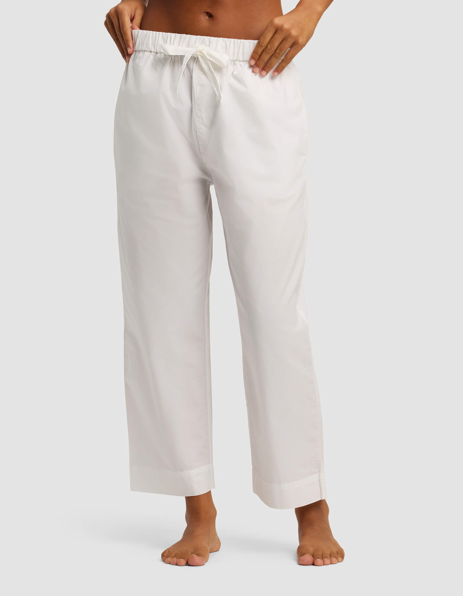 A woman stands barefoot against a plain light background, wearing Cozy Earth's Women's Sutton Short Sleeve & Pant Pajama Set with loose-fitting white drawstring pants cropped at the ankles. The upper body is partially out of frame.