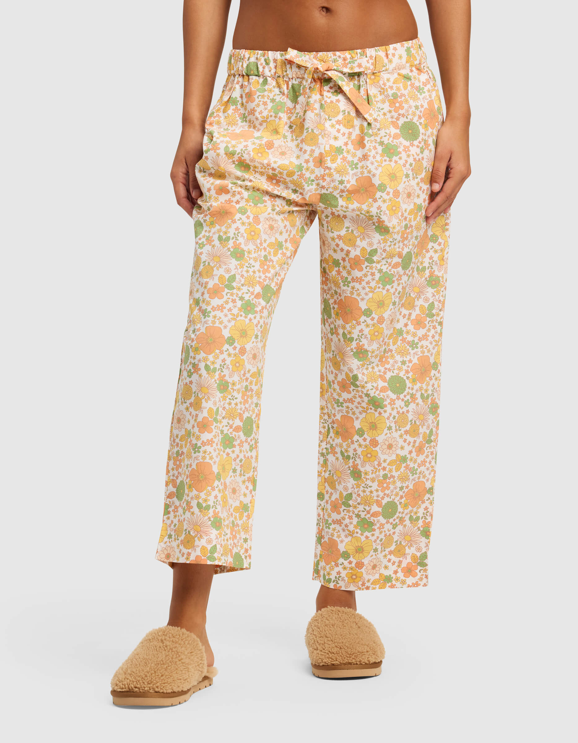 A model wears Cozy Earth’s Women’s Sutton Cropped Pajama Pant, featuring a pastel floral print, paired with fuzzy beige slippers; photo shows the look from waist to feet against a plain light background.