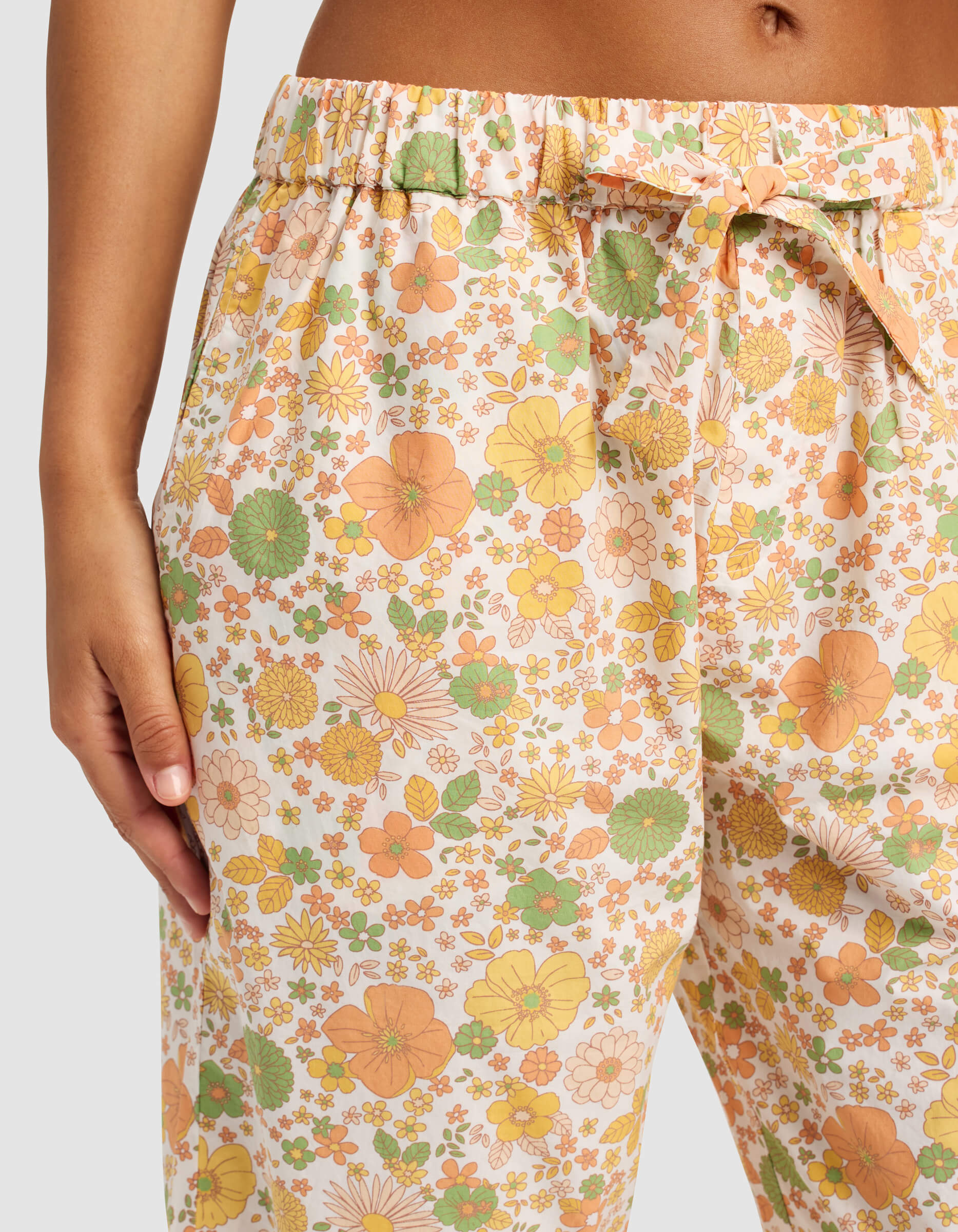 A person is wearing Cozy Earth's Women's Sutton Cropped Pajama Pant, featuring an orange, green, and yellow floral pattern, light-colored fabric, a waist tie, and pockets.