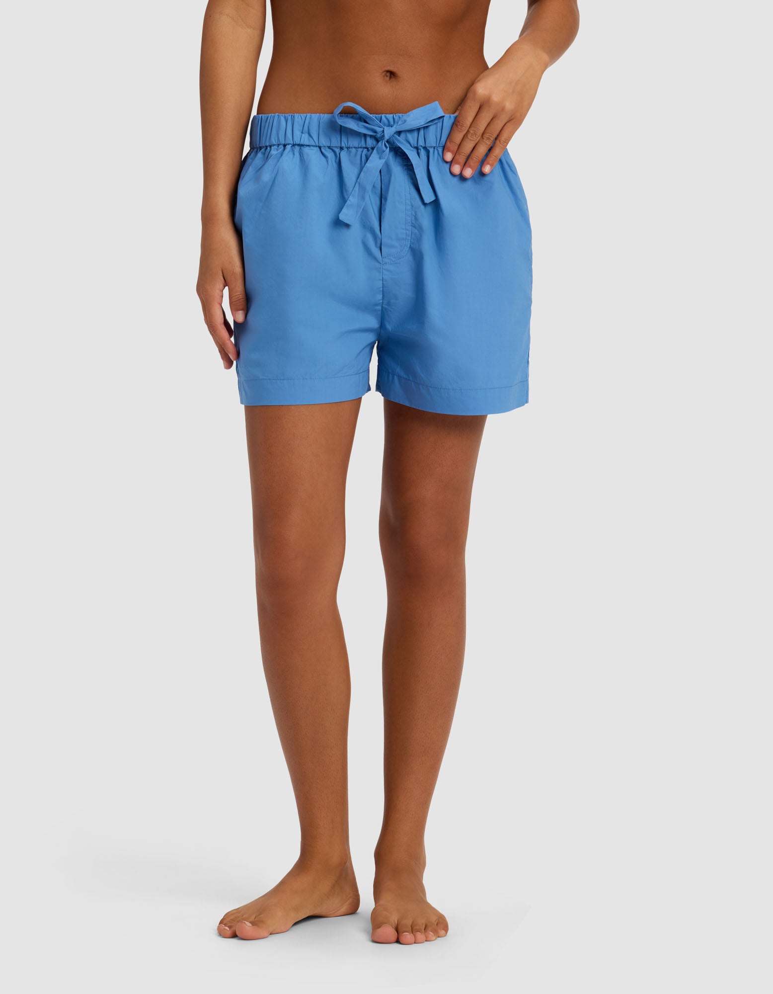 A person stands barefoot against a plain white background, wearing Cozy Earth's Women's Sutton Pajama Short in light blue with a tied drawstring bow at the waist.
