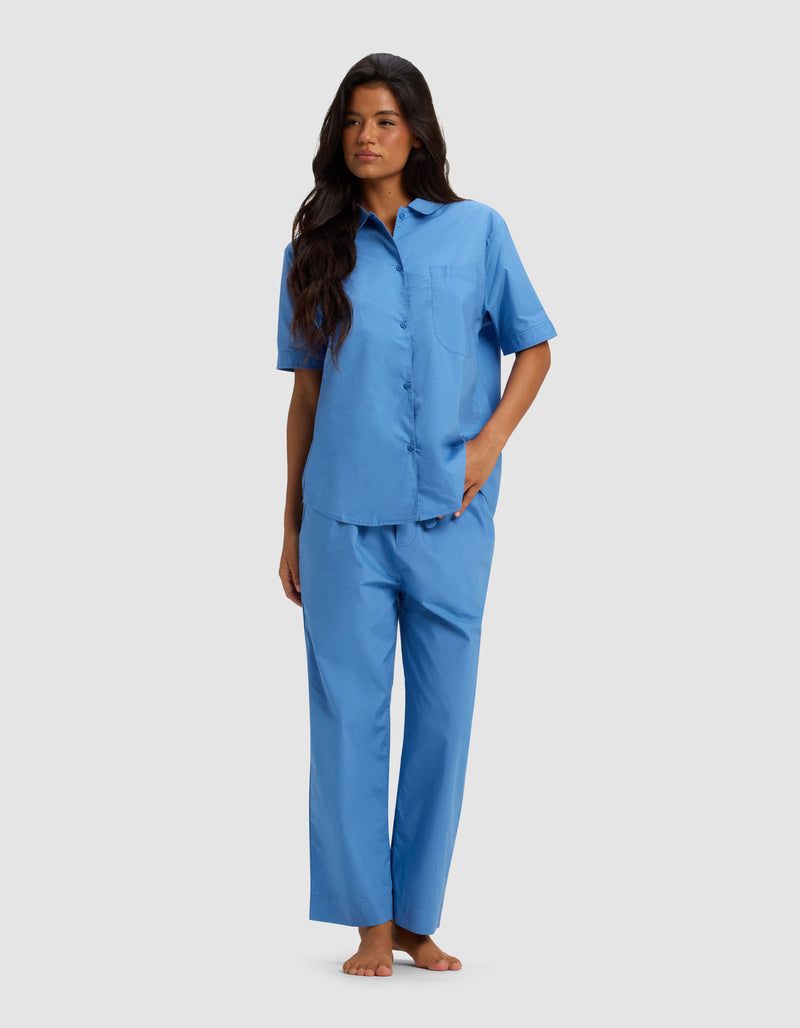 A woman with long dark hair stands barefoot against a plain background, wearing the Cozy Earth Women's Sutton Short Sleeve & Cropped Pant Pajama Set in light blue, with one hand in her pocket and gazing slightly to the side.