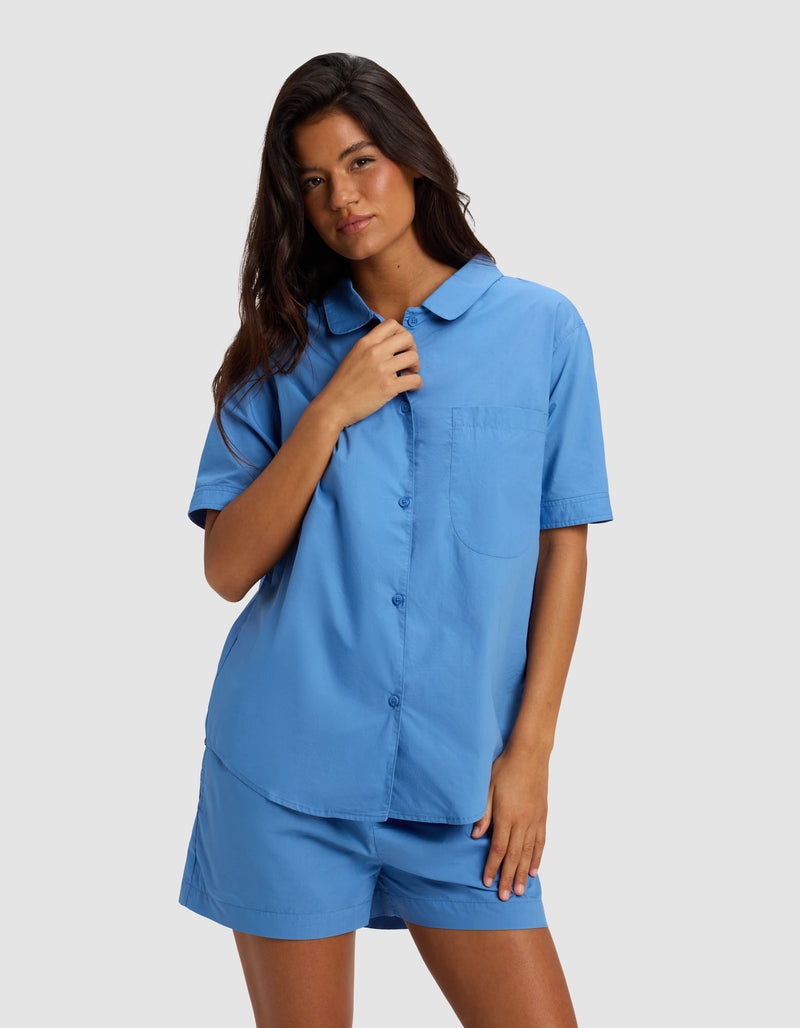 A woman with long brown hair wears the Cozy Earth Women's Sutton Short Sleeve Pajama Top in light blue, standing against a plain light gray background. She touches her collar with one hand and rests the other by her side.