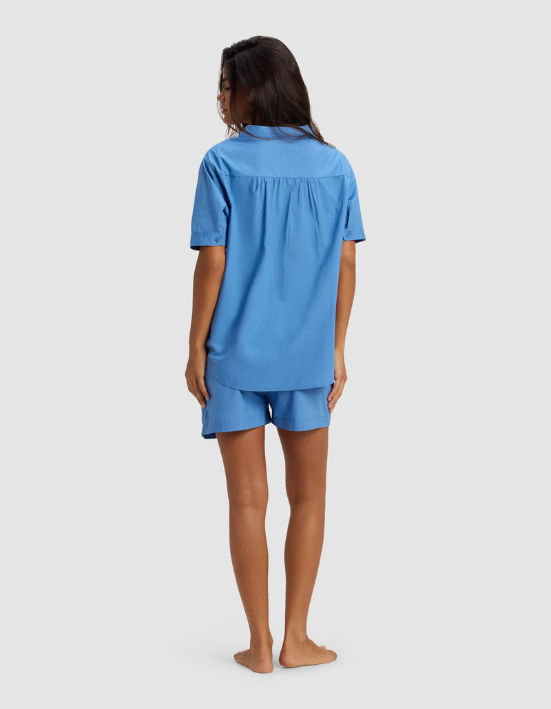 A woman stands barefoot, facing away, wearing the Cozy Earth Women's Sutton Short Sleeve Pajama Set in light blue, featuring a loose-fit shirt and matching shorts against a plain light gray background.