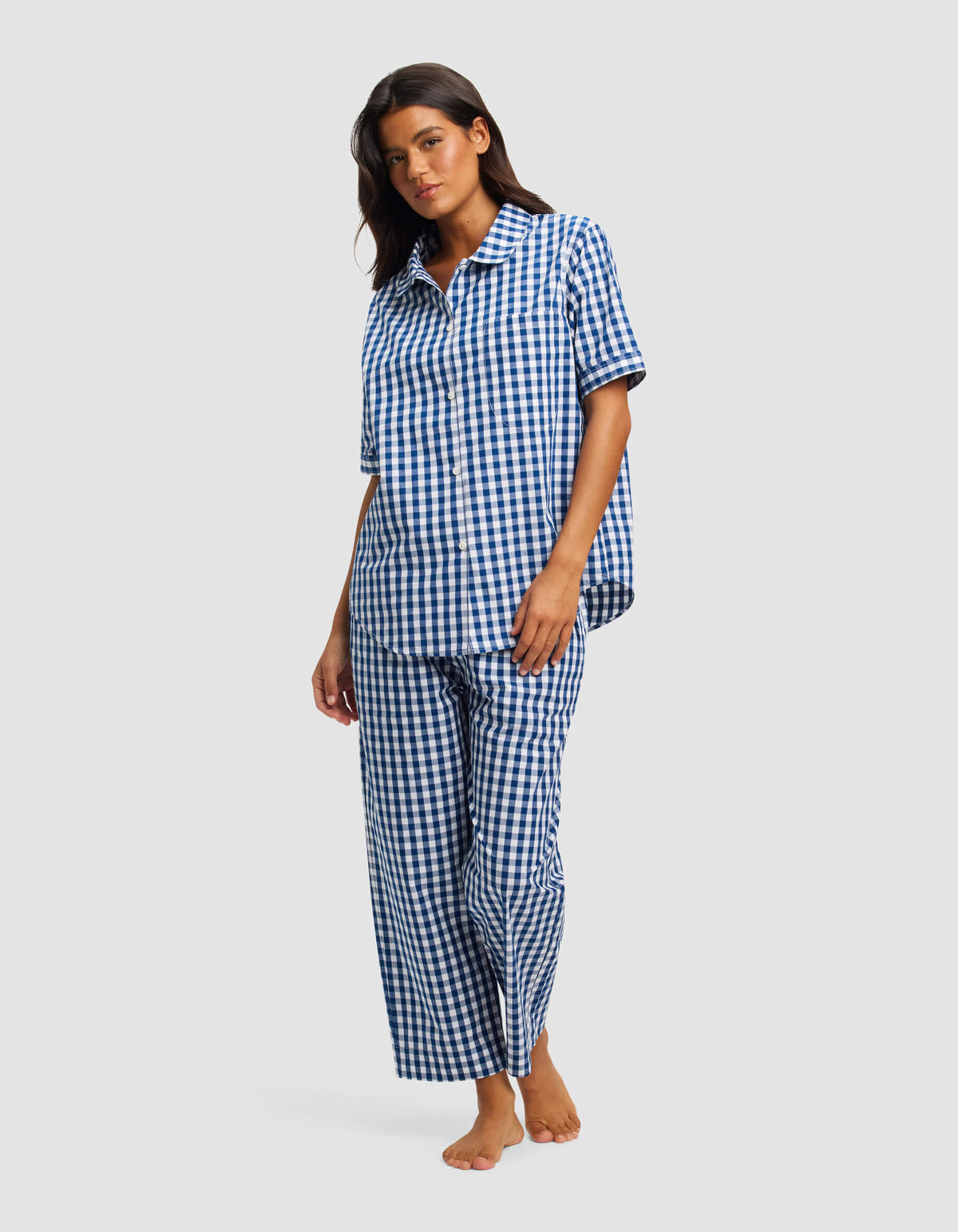 A woman stands barefoot against a light gray background, wearing the Cozy Earth Women's Sutton Short Sleeve & Pant Pajama Set in a blue and white checkered pattern.|Color:Pacific Gingham