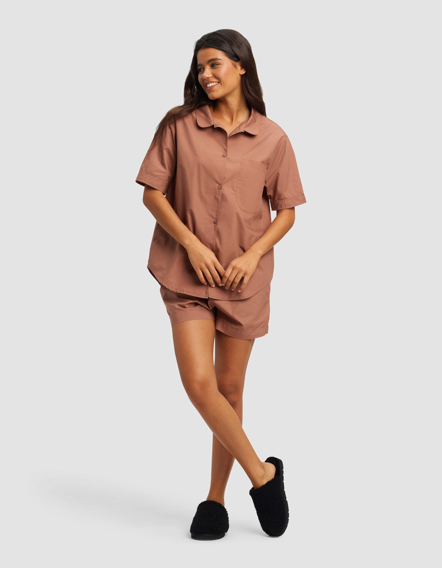 A woman smiles in the Cozy Earth Women's Sutton Short Sleeve Pajama Set—featuring a brown button-up top with matching shorts and black slippers—against a plain light gray background.