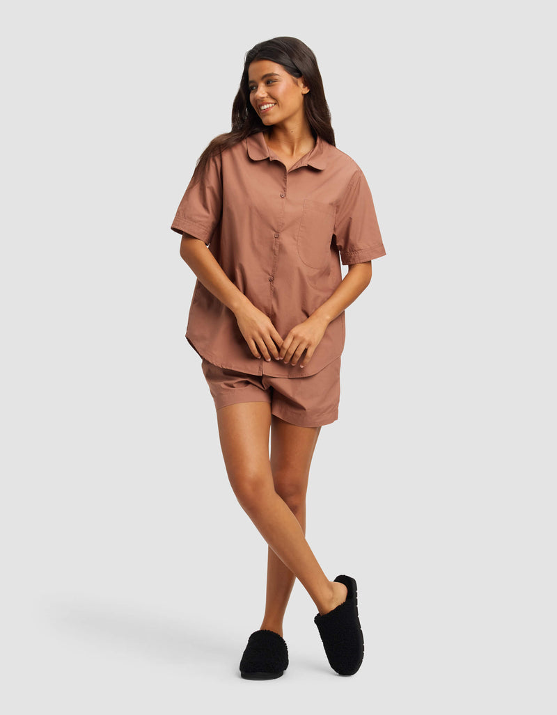 A woman smiles in the Cozy Earth Women's Sutton Short Sleeve Pajama Set—featuring a brown button-up top with matching shorts and black slippers—against a plain light gray background.