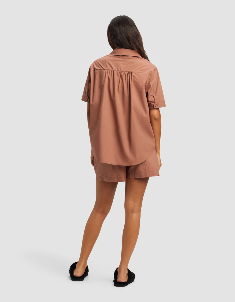 A woman faces away, wearing the Cozy Earth Women's Sutton Short Sleeve Pajama Set in light brown and black slippers. Her wavy hair is down against a plain white background.